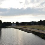 View from a bridge over a river. The river passes into the distance to the left, then curves around to the right, arcing around a large lawn with a small white pavilion in the distance. The horizon on all sides is blocked by trees. The sun is low in the sky, and shines from behind a large cloud.
