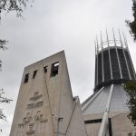 A modern cathedral building. On the left is a stone frontage with a carved pattern of crosses, and four bells in notches near the top. The frontage is separate from the main building, which has a main roof which is a pyramid based with a regular polygon of many sides, and on top of this is a cylinder glazed with stained glass, on top of which are a series of crown-like spires. There is a grey sky behind, and there are trees on either side.