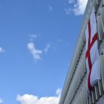 Looking up at a blue sky dotted with clouds. Across the right-hand side of the image is a three-storey car park, concrete, with twisted cream-coloured fins along each balcony. A large England flag hangs from the top storey to the bottom. There are a couple of silhouettes of chimneys at bottom right.