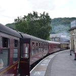 Crimson-coloured old rail carriages along a curved platform, with steam from a steam engine in the distance. There are antique lampposts and stone buildings on the platform, and “MIND THE GAP” written in white paint along the edge. In the background is a wooded hill, with some mill buildings beneath.