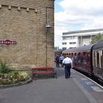 Old crimson-coloured rail carriages along a railway platform, with “MIND THE GAP” in white paint along the edge. On the left is a large stone building with a plaque on it (rectangular, with arc protrusions at top and bottom. The sign is crimson, with a white border and white text reading “KEIGHLEY”. Below the stone wall is a wooden bench, painted crimson and black, and to the left of that a flower bed. In the background is a four-storey modern white building.
