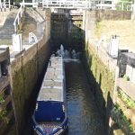 A canal boat in a lock, seen from above the lock’s lower gates, which are open. The upper gates are closed, and there is another, higher, set of gates beyond them, also closed. There are steps and bridges around the locks. A mill building sits alongside the canal to the right, just trees to the left.