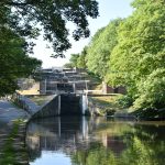 A view of a staircase lock of five rises, from the bottom. The lowest lock is reflected in the canal, as is an outbuilding and as are some of the trees with surround the canal. The view is from the towpath.