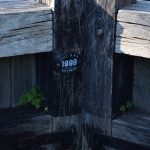 A detail of the join between two lock gates. The gates are of dark, unpainted wood. There are small plants growing from some of the cracks between the beams making up the gates. At the lip of the left-hand gate is a black metal plaque, with white writing. The plaque is oval-shaped, and along the top of the oval is “BUILT B W B”, across the middle (in very large text) “1999”, and along the bottom of the oval “NORTHWICH”.