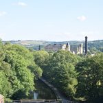 A view from high above a canal. The canal is surrounded by trees, above which can be seen mill buildings of yellow stone, with a black-painted chimney, and further in the distance steep rural-looking hills.