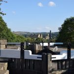 A canal lock, with two gates visible, seen from behind the higher one. The gates are black, with white handles, and the canal is lined with stone towpaths on either side, with trees behind them (and behind a stone wall on the left). Above the lock can be seen mill buildings of yellow stone, with a black-painted chimney, and further in the distance steep rural-looking hills.