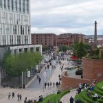 A modern shopping centre, open-air, with a staircase made of artificial grass steps, next to a red-stone wall. Beneath is a long precinct with many pedestrians, and to the left a modern hotel building, white with many high windows. In the background are red-brick high dock buildings, one with a red-painted portico, and a red-brick chimney.