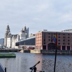 A view across a dock, with red-brick buildings around the edge. There are no boats in the dock; it is large and square. There is a colonnade around the dock, supported by red pillars (which are all that can be seen at the extreme left. Through a gap in the dock buildings on the far side to the left are more stone buildings, and part of one made of glass. One of the stone buildings has a central dome in a tower. Behind that is a building with two towers, one of which has a clock; both towers are topped with a small copper bird.