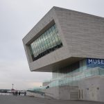 A museumL the text “MUSEUM OF LIV…” is visible on its side. The lower part of the museum building is obscured by a zig-zag ramp with glass railings. Above that is a metal box (irregularly-shaped with sloping walls), one side of which has a large window area made of smaller panes, with “IMAGINE PEACE” visible in the windows. The dark sky is visible around most of the building. The River Mersey is visible in the background, with a few people walking along it.