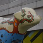 The front of a statue of a lamb, with part of its banana-shaped tail obscured. The lamb is painted with a design with a sea and fish on the lower half, and an abstract tree with birds (some on the water, some in the tree) on top and on its head. Behind it is a metal building.