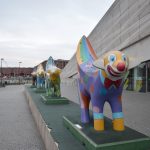 A parade of four painted lambs whose tails turn into bananas. The front one is painted like a clown; the designs of the ones behind are less visible. To the right is a metal building, with a glass wall int he lower part. Behind the precinct on which the Superlambananas stand is a waterway, and behind that are red-brick dock buildings.