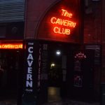 A darkened building entrance leading into a club. There are two neon signs, red, reading “The CAVERN CLUB”, one above the door and one to its left, and several other white-on-black signs with the same title. The club door is open, and a couple of people are around.