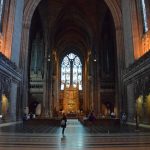 Looking up the nave of a red-stone cathedral, with a very high gothic-arched ceiling. There are several tourists milling around, and some wooden pews, which do not start until near the background. On the far wall is a stained glass window, which mostly appears white. Partway down the nave are the edges of the transepts, with organ pipes on the top of their walls and small gothic arches beneath them. A high altar is also visible at the very far end, with a wooden altar.