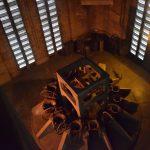 Looking down inside a large red-brick chamber, dimly lit, towards a set of bells arranged in a circle around a large wooden frame. Two walls of the chamber are visible, with horizontal slit windows, in two groups of two columns on each wall.