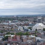 Looking from a high place across city development, which starts out as a city centre and becoming suburban, though there are a couple of football stadia (one larger than the other) near the skyline. The Metropolitan Cathedral (a conical building, with a stained-glass cylinder on top (itself topped by crown-like spires) is prominent in the middle-distance to the right.