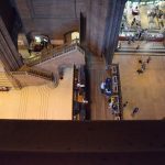 Looking down from a balcony into the nave of a cathedral. There is a white floor to the left and a light stone floor to the right, with a wooden section in between. One wall has a staircase doubling back and leading to the wooden portion. Through an archway on the far wall to the right is a brightly-lit gift shop. The balcony from which the photo is taken takes up the lower third of the picture.