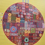 An abstract mural on a tiled wall. The background is yellow, with a red circle on it. To the left and right are five rows of tiny circles of various colours, and three vertical grey lines are “attached” to the top of the circle. The red circle contains patterns, mostly circular or rounded, of various colours and shapes. To the right of the wall, two landings can be seen with railings.