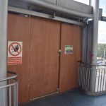 A boarded-up passageway from a blue-floored landing with metal railings. Behind the railings is a glass wall with metal struts, and through one of the windows can be seen a dual carriageway and a church. On the boarding is a “SITE SAFETY” notice, and a sign with a human figure with an outstretched hand (black) crossed through in red in a red circle, with white-on-red lettering beneath reading “No unauthorised persons allowed beyond this point”.