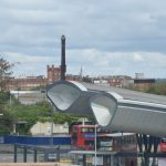 An elevated view across a town. In the foreground is a bus station, whose roof is made of two silvery tubes, the cross-section of which is a triangle with a curved top. There is a.modern building on the left. In the background is a red-brick factory with a tall dark chimney. The factory has cut-out letters supported on its roof by white metal struts, spelling out “HORLICKS”.
