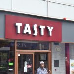 A coffee shop in a modern building. The sign is white-on-burgundy, and reads “TASTY”, in the same font as the Costa logo (including the S). A man stands in the doorway, with one of the double doors ajar. WHSmith, a phone booth, and two youths are reflected in the window.