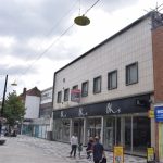 A pedestrianised shopping street. The visible shops are an unidentified boarded-up unit, an empty BHS store (with an “ALL ENQUIRIES” sign suspended from its upper, tiled, wall), and “MOBILE KING”, a computer and mobile repair shop. The sky is dark and brooding. The street is moderately busy.