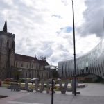 A church, made of mottled dark-and-light stone, with a red roof and red spire on its castellated tower. In front of it is a garden, and to the right a modern, curved building, with a glass wall at the end of the curve. In front of the garden is a sign made of cut-out letters reading “THE CURVE”, and in front of that traffic lights.