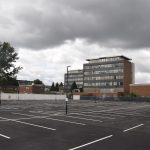 An empty car-park, with fresh-painted white lines on its tarmac. The far edges of the car park are hoarding. Behind the hoardings are various industrial buildings, including a six-storey office building with two plate-glass walls and two brick ones. Above the scene is a dark cloud.