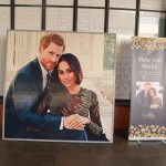 A large picture of Prince Harry and Meghan Markle, sitting next to each other and holding hands, made out of Lego bricks. A free-standing banner reads “Make your Markle” next to it. The mural is in a frame, leaning against a papered-over shop window (with line drawings of bricks and silhouettes of people on the paper). There is a bicycle wheel visible on the far right. There are square concrete paving slabs making up the floor.