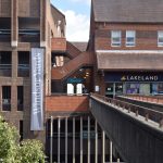 View from one side of a brutalist footbridge high scores a road. The footbridge is narrow, and leads to a shopping centre, with a Lakeland shop visible. The shopping centre is brick, with a row of windows above the shop fronts. To the left is an outdoor staircase above a passageway, and to the left (and below the shops) a multi-storey car park, upon which a vertically hung white-on-lilac banner reads “Windsor Yards”. A few cars, along with a tree, are visible on the street below the footbridge.