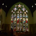 A large, floor-to-ceiling, stained glass church window, featuring gothic arches in the stonework, and various brightly-coloured human figures. A person is silhouetted in front of it, as is an altar and altar rail. Some pews are just visible. The wall is white, appearing yellow in the low light, and the ceiling is invisible, save for a few white spotlights.