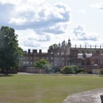 A view from the top of a wall over a playing field, towards the buildings of Eton College. These are mostly of red brick, several storeys, with castellated tops of walls; there are three trees to the left. To the right, above the red-brick building, is a light-stone chapel, with the top of the long wall visible, also castellated, with small spires rising between every gothic window.