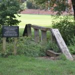 Over a stream from a field passes a wooden beam, with ramps up to it of the same width. There are two (deliberate) breaks in the beam, which looks slightly rickety. On the far side is a playing field, with a brick walls and a large red-brick building at the other side. Next to the beam, on the field, is a sign that reads, in white-on-black text: “THIS EQUIPMENT IS FOR USE BY THE STAFF & BOYS OF ETON COLLEGE ONLY UNDER THE SUPERVISION OF A QUALIFIED PERSON. MEMBERS OF THE PUBLIC ARE KINDLY ASKED NOT TO USE OR TAMPER WITH THE EQUIPMENT. FOR HYGIENE REASONS DOG OWNERS SHOULD NOT ALLOWED THEIR ANIMALS TO FOUL THE EQUIPMENT OR AREA.”
