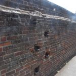 A brick wall, from foreground on the left to background on the right. The wall is high, and set into into are a series of iron footholds and a hand-hold allowing people to climb to the top.
