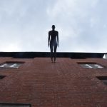 A metal sculpture, life-size, of a man’s standing form, attached by the feet to a wall so that it protrudes horizontally outwards. The wall is brick, and three storeys of window can be seen. The statue is silhouetted against a cloudy sky.