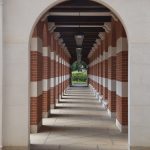 A view along a colonnade, with arched entrances in white stone at either end and square brick-and-white-stone columns along the edges. The roof is of dark wood, supported by white stone brackets. A hedge and garden can be seen at the far end. The columns make a repeating pattern of shadows along the concrete-paved floor.