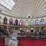 A view inside an oval-shaped building with an ellipsoid roof (supported only by the external walls and its own structure; the whole thing is painted cream, and there is a skylight). There is a bacon, with brightly-colour shop doors under arches, and more muted-coloured doors on the main floor. There is a hole in the floor down to the basement in the foreground.