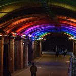 A brick tunnel with a curved roof, and a road along the bottom. There are rainbow-coloured lights lighting the ceiling. The road is empty of cars, but there are various people walking up and down it.