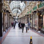 A lavishly-decorated Victorian arcade, with uniform shop fronts beneath a narrow balcony with cream tiling and green ornate low railings, the whole being topped by an arched roof with skylights. The arcade is busy, and has globe-shaped lights along the centre.
