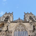 Looking up at the side of a Cathedral. There are two towers on either side of the central wall, with gothic arched windows in both. There are is a small turret on each corner, and in the middle of each side, of each tower. The central window, in between gothic decoration, has a heart-shaped pattern in the stonework. Looking up, the sky is blue, flecked with clouds.