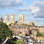 A huge cathedral, with two west towers and a central tower, is visible on the skyline of a view from some height. In front are brick buildings, and a road crossing a white bridge. On the left of the picture, in front of some trees, are two parallel sets of white railings that arch over a bridge, marking the edges of the wall-top path.