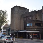 An art-deco brick cinema with lettering on the wall reading “ODEON”; the letters above the entrance say “EVERYMAN”, with a sign with moveable letters beneath (too small to read). It is on the opposite side of a busy road, at a traffic light crossing.