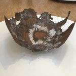 A piece of metal, with salt deposits in parts. It is shaped like a cut-out section of an inside-out globe, and is the portion corresponding to the Indian Ocean, so the outlines of Madagascar, India and Sri Lanka can be spotted, among others. It sits on a while circular plinth on a wooden floor; there is a similar plinth with a similar piece of metal in the background. Also visible (partly off-frame) is a slice of a tree on a rectangular white plinth.