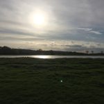 Looking across a field, which is partly flooded, forming a lake. In the distance are a line of trees, and hills, beneath a clouded sky. The sun shines at the camera through the clouds, and is reflected in the lake.