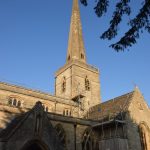 A cruciform church, made of Cotswold stone, with a tall spire atop a central tower. The leaves of a tree overhang.