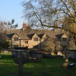 A long Cotswold stone house seen across a turfed graveyard. There is a bare tree to the right.
