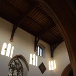 Three light patches, shaped the windows opposite, on a whitewashed church interior wall. There is a high plain window and a lower Gothic stained-glass window. The roof is bare wood.
