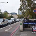 Looking from a curb along a busy road, with traffic built up. There are thick trees on either side of the road, and on the horizon a cluster of tall buildings. On the pavement are railings with Underground roundels on poles, and blue signs reading “REGENT’S PARK STATION” in white on blue. The railings guard steps underground.