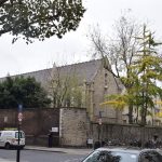 Looking diagonally across a T-junction towards a stone building with a high hipped roof and few windows, enclosed in a high brick-and-stone wall. Other buildings inside the wall are visible. The road is quiet, except for one person crossing the junction, though various parked cars, bikes and motorbikes can be seen.