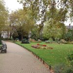 A gaden in a park, with a path with low railings leading away from the camera. On either side are lawns; on the left with tall cone-shaped hedges, on the right with small flower beds, one with red flowers. Various trees overhang the scene. A row of red terraced houses is visible outside the park.
