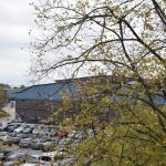Looking from a height down at a factory and its car park. The factory (red brick two storeys, with a blue corrugated section of wall at roof-height) is mostly hidden behind a tree on the right, whose yellowish leaves are starting to fall. The car park is full.